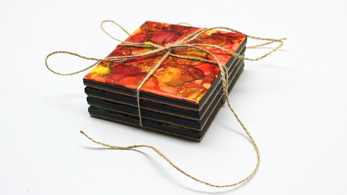 A stack of four ceramic tile coasters marbled in red, orange and gold alcohol ink with black edges, tied with jute twine