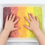 Child's hands on top of a tray of rainbow sand.
