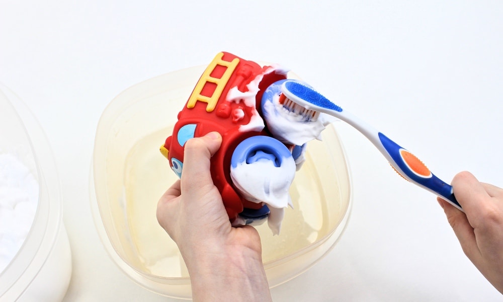 Child's hands washing shaving cream off a toy car with a toothbrush.