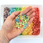 Hand holding up rainbow-coloured oats on top of a bin of rainbow-coloured oats.