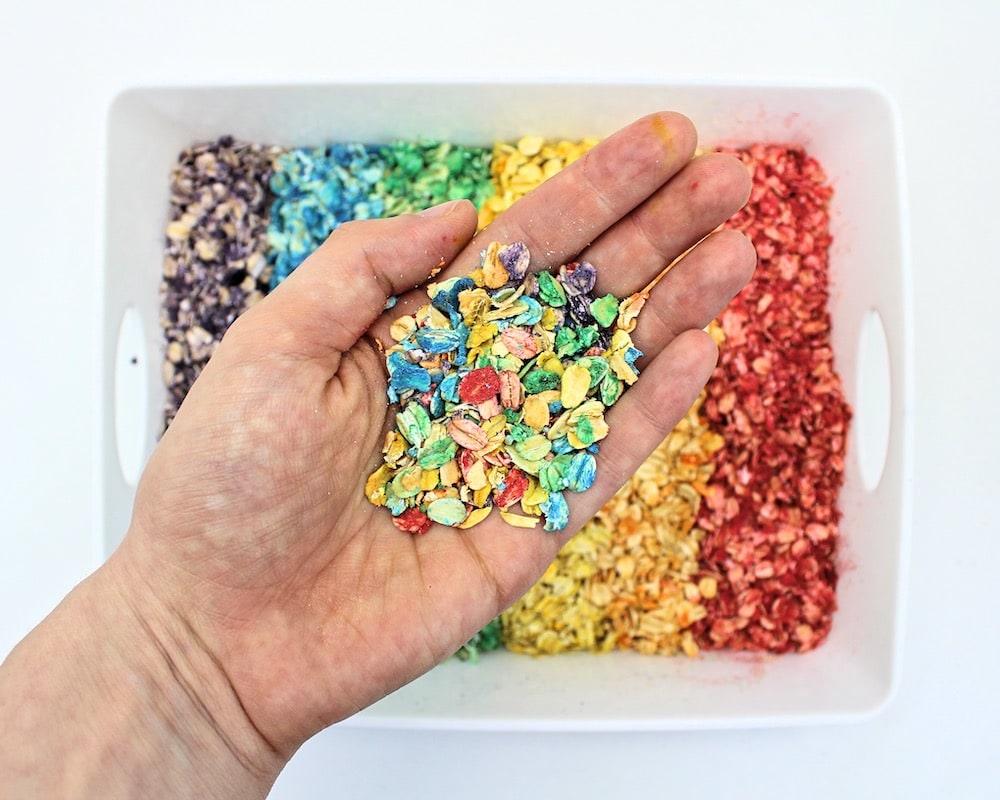 Hand holding up rainbow-coloured oats on top of a bin of rainbow-coloured oats.