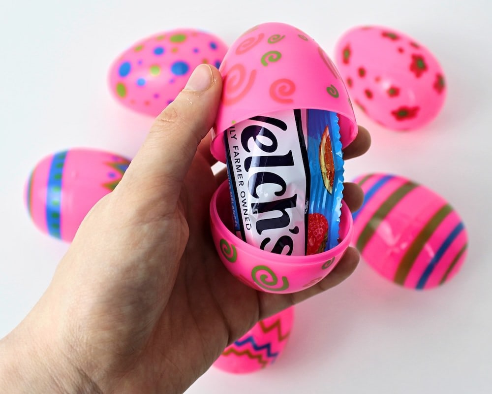 Hand holding up a pink egg containing a pack of Welch's fruit snacks, with more pink eggs in the background.