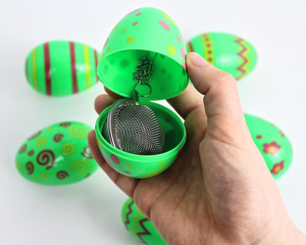 Hand holding up a green egg containing a tea infuser, with more green eggs in the background.