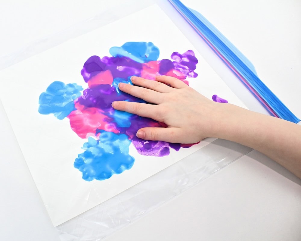 Child's hand smushing paint around inside a Ziploc bag.