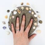 Hand pressing down on a ball of play dough surrounded by seashells.