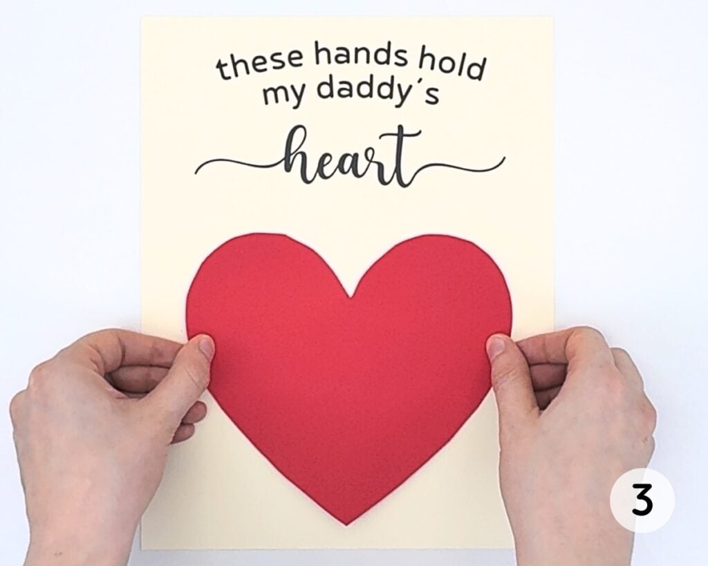 Hands pressing down red heart onto beige cardstock.