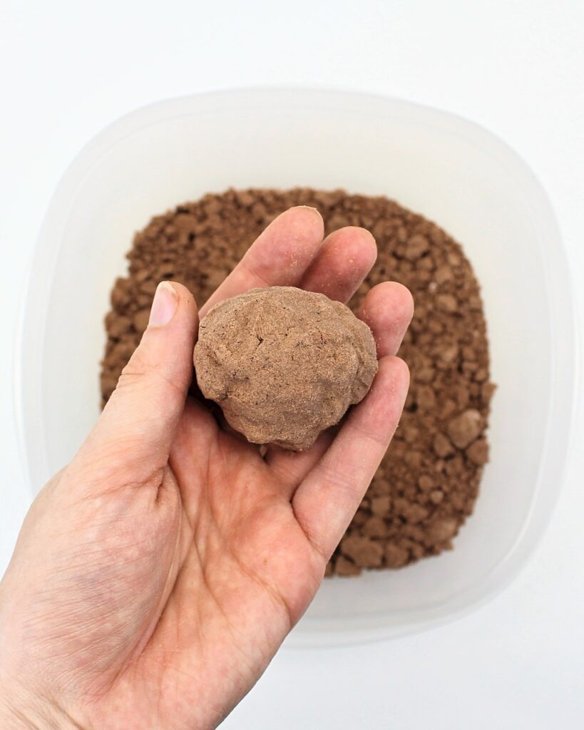 Hand holding up a brown ball of sand on top of a bin filled with brown sand.
