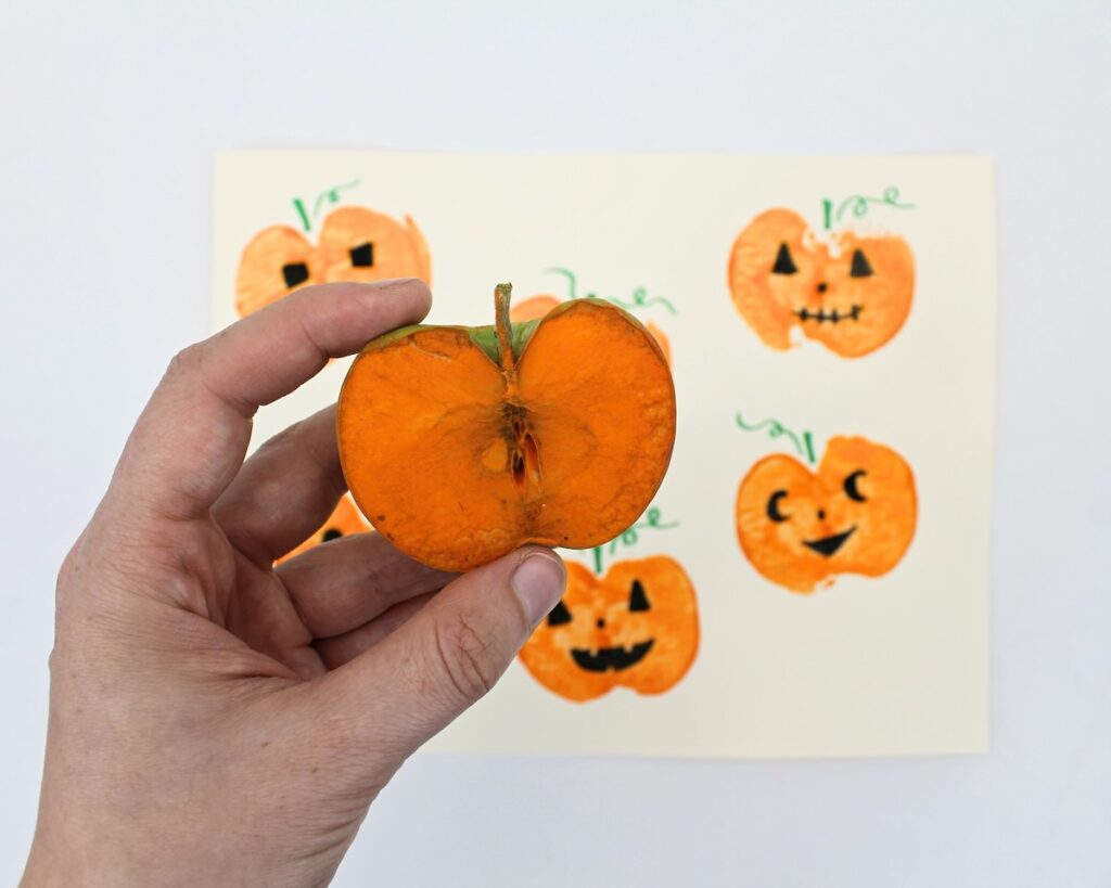 Hand holding up an apple half with orange paint on top of the finished jack-o'-lantern craft.