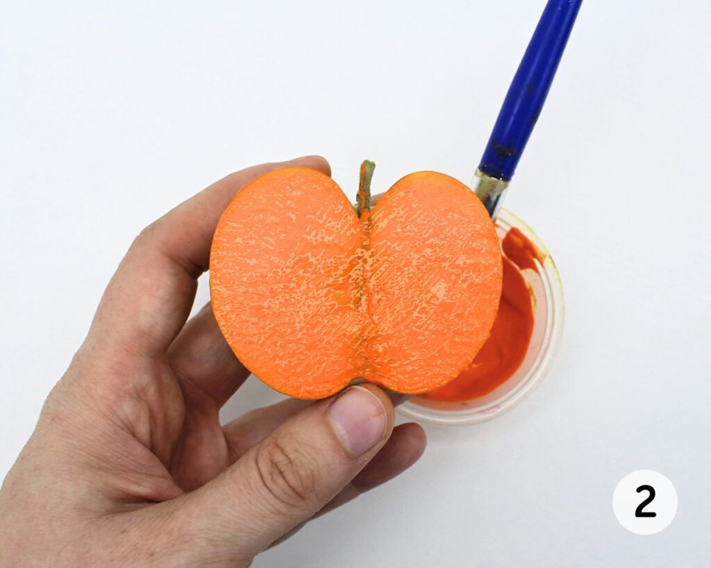Hand holding up an apple half covered with orange paint.