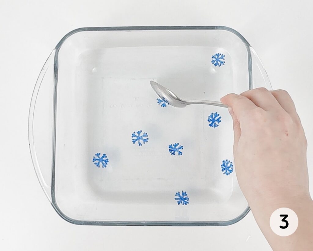 Hand dipping a spoon into a dish of water with snowflake drawings floating inside.