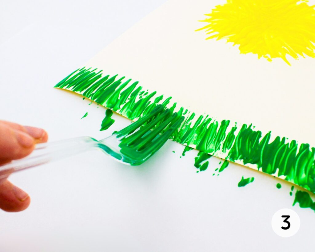 Fork covered with green paint, painting blades of grass at the bottom of a sheet of cardstock.
